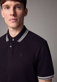 Fekete polo póló csíkos gallérral, két gombbal és hímzett logóval. Texturált anyagból készült, testhezálló fazonnal.