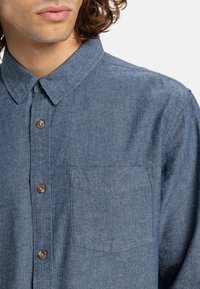 Denim shirt with a button-up front, collared neckline, and a single chest pocket. Material is textured, in a medium blue hue with brown buttons.