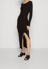 Robe longue noire à manches longues côtelées avec un col et un ourlet de couleur crème, dotée d'une fente latérale et portée avec des mules noires à talon bloc.