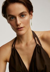 Persona joven con cabello corto castaño, vistiendo un top halter oscuro y un pendiente colgante con detalles en verde y dorado.