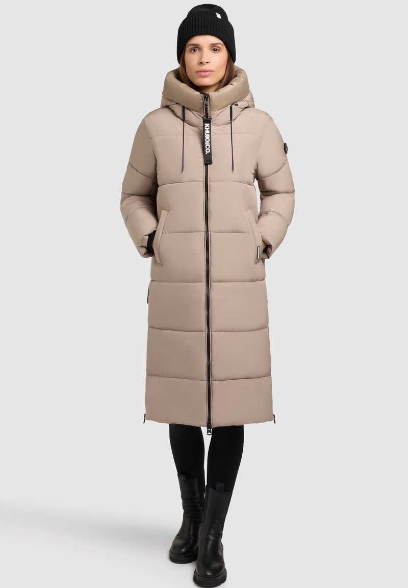 Beige puffer coat with a hood, featuring a front zipper, side pockets, and a quilted design. Paired with a black knit hat and black boots.