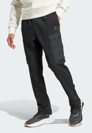 Pantalons de jogging noirs avec une texture lisse, dotés de poches cargo en tissu contrasté, associés à des chaussures de sport grises.