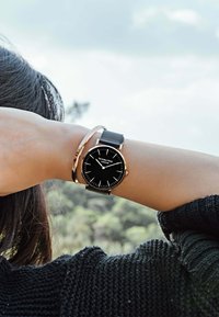 Montre noire avec un cadran rond et des accents dorés, dotée d'un bracelet en maille noire et d'un bracelet fin noir, le tout sur fond naturel.