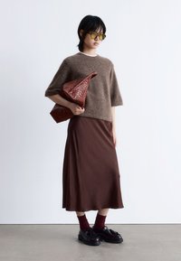 Braun gestrickter Pullover, braune satin Midi-Rock, komplizierte bordeauxrote gewebte Handtasche, gelbe Sonnenbrille, schwarze Schuhe mit dicken Sohlen und Akzenten.