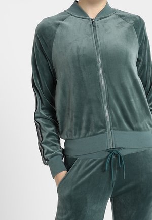 Woman wearing dark green velour zip-up jacket and matching drawstring pants with black stripes on sleeves.