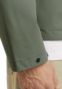 Maniche di giacca verde oliva con una texture liscia, caratterizzate da un polsino curvo e un dettaglio con bottone nero. Lo strato interno è bianco.