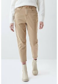 Pantalones de algodón beige con un corte relajado, que cuentan con dos bolsillos frontales, cierre de botón y dobladillos recortados, combinados con botas de tobillo de color claro.