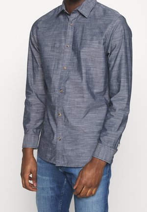 Homme portant une chemise bleue à manches longues boutonnée et un jean bleu, les mains détendues le long du corps.