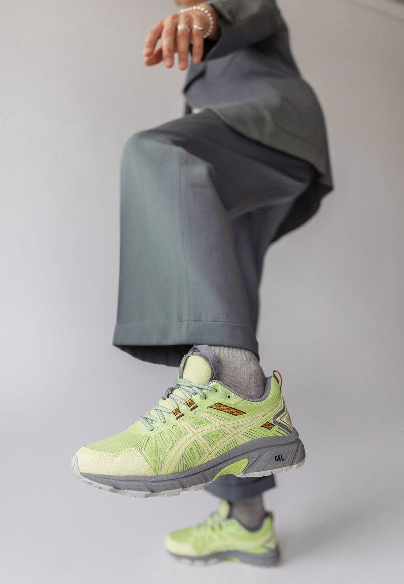 Personne en pantalon gris levant le pied chaussé d'une chaussure de course vert citron et grise avec des chaussettes grises sur fond uni.