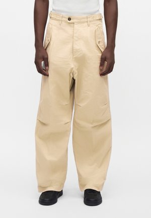 Personne portant un pantalon beige à jambes larges avec des poches latérales boutonnées et des chaussures noires sur fond blanc uni.