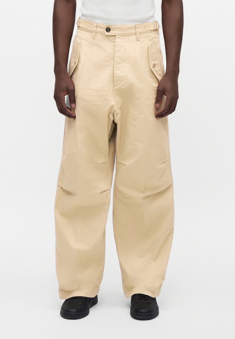 Personne portant un pantalon beige à jambes larges avec des poches latérales boutonnées et des chaussures noires sur fond blanc uni.