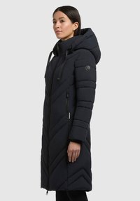 Lang navy puffer jakke med et chevronmønster, hætte, frontlynlås og sidelommer; har tekstureret stof og logodetalje på ærmet.