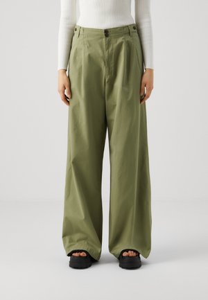 Vrouw draagt olijfgroene broek met wijde pijpen, wit geribd shirt met lange mouwen en zwarte platformsandalen met zichtbare tenen.