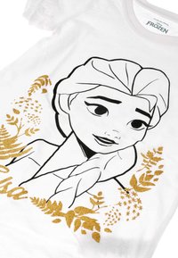 Witte T-shirt met een illustratie van het gezicht van een personage in zwarte omtrek, versierd met gouden glinsterende bladpatronen en details.
