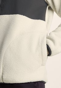 Cream-colored fleece jacket with gray fabric shoulder section, featuring a front pocket and textured material for warmth.