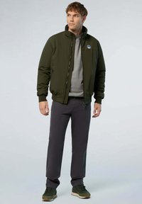 Giacca bomber verde oliva con colletto alto, zip frontale e tasche laterali, indossata sopra un maglione grigio e pantaloni scuri, con sneakers verdi.