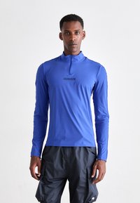 Hosszú ujjú, cipzáras nyakú sportpóló élénk kék színben, fekete "Calvin Klein" logóval a mellkasán. Könnyű, rugalmas anyagból készült.