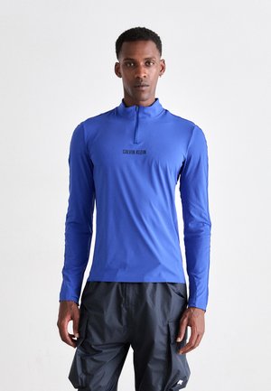 Pitkähihainen, vetoketjullinen urheilupaita kirkkaansinisenä, jossa musta "Calvin Klein" -logo rinnassa. Valmistettu kevyestä, joustavasta kankaasta.