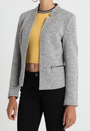 Kvinde iført en grå tekstureret lynlåsjakke over en gul cropped top og sorte bukser, vist fra hals til hofter.