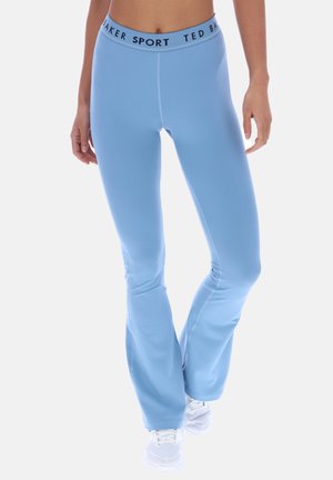 Kvinne iført lyseblå vid treningstights med "Ted Baker Sport" i linningen og hvite joggesko, stående mot hvit bakgrunn.