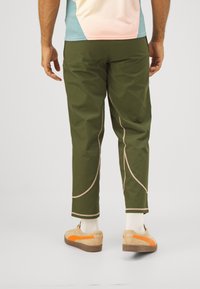 Grüne, leichte Hosen mit geschwungenen beigen Nähten, in cropped Länge und mit glatter Textur, kombiniert mit beigen Sneakers mit orangenen Akzenten.