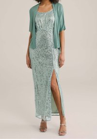 Dlouhé mintově zelené šaty pokryté flitry s vysokým rozparkem na stehně, doplněné o odpovídající otevřený šál a stříbrné sandály.