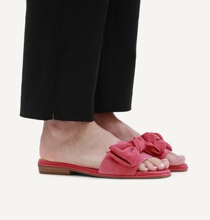 Feet wearing open-toe pink suede slide sandals with large decorative bows, paired with black cropped pants on a white background.