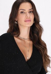 Schwarzer Pullover mit glitzernden Akzenten; geschichtete silberne Halsketten, eine mit einem runden Anhänger; Model mit welligem braunem Haar vor neutralem Hintergrund.