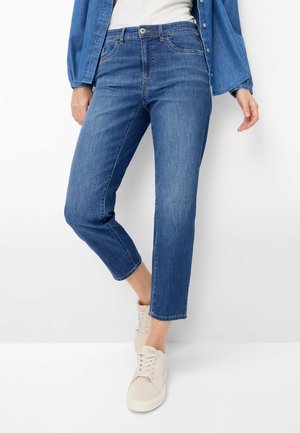 Frau trägt mittelhohe, blaue, verkürzte Jeans, blaue Jeansjacke, weißes Oberteil und beige Schnürsneaker, stehend vor einem schlichten weißen Hintergrund.