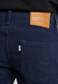 Mörkblå manchesterbyxor med en lädermärkning på baksidan av midjebandet och en synlig bakficka. Har horisontell ribbad textur.