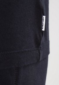 Detailní záběr na černý šev látky s bílou značkovou cedulkou Converse přišitou na okraji.