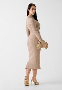 Sukienka dzianinowa w jasnobeżowym kolorze z fakturą prążkowania i dopasowanym krojem, zestawiona ze złotą kopertówką oraz sandałami na wysokim obcasie z paskami.