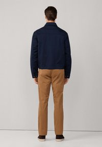 Giacca blu navy con colletto classico, mostrata da dietro, abbinata a pantaloni khaki e scarpe marroni scure. Tessuto liscio, design minimalista.