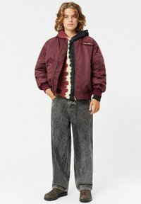 Maroonin värinen bomber-takki, jossa on ribbed-hihansuut, mustan ja tie-dye-hupparin päällä, yhdistettynä haalistuneisiin mustiin leveisiin farkkuihin ja ruskeisiin kenkiin.