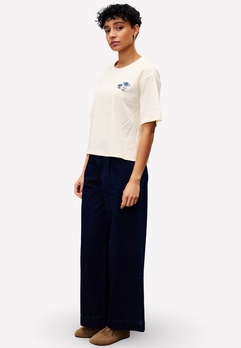 Vrouw met kort krullend haar draagt een crèmekleurig T-shirt met een klein bloemenpatroon, losse donkerblauwe wijde broek en bruine schoenen, en staat zijwaarts.