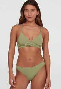 Femme aux cheveux longs portant un bikini vert olive clair avec un haut style drapé et un bas assorti, se tenant devant un fond gris clair uni.