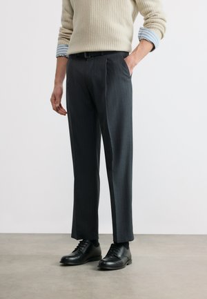 Άνδρας που φοράει μαύρο παντελόνι φόρμας με ρίγες, μαύρα δερμάτινα παπούτσια, μπεζ πουλόβερ πλεκτό με μανίκια μπλε ριγέ πουκάμισου τυλιγμένα, στέκεται με το ένα χέρι στην τσέπη.