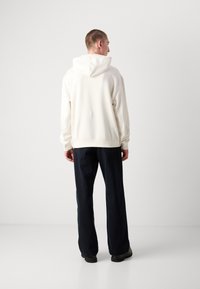 Sudadera blanca con capucha, con un corte holgado y puños acanalados, combinada con pantalones de pierna ancha azul oscuro y zapatos negros, vista desde atrás.