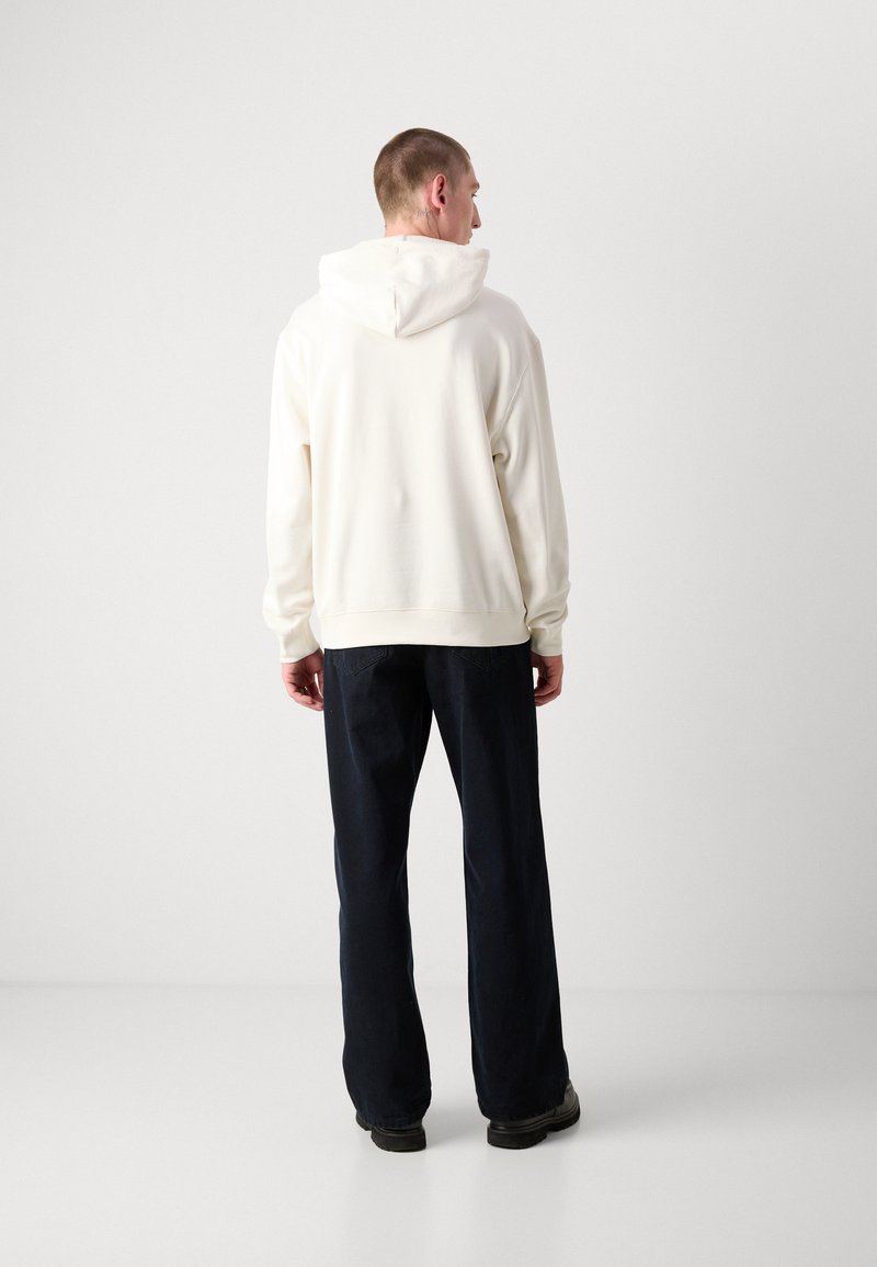 Felpa bianca con cappuccio, dal taglio rilassato e con polsini a costine, abbinata a pantaloni a gamba larga blu scuro e scarpe nere, vista da dietro.