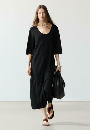Donna che cammina con un vestito midi nero con maniche a gomito, porta una borsa nera e indossa sandali marroni.