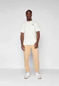 T-shirt de manga curta verde claro com um bolso no peito, combinado com calças de moletão bege e ténis brancos. Design simples, ajuste casual.