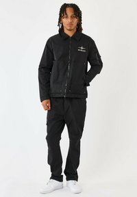 Giacca nera con chiusura a zip, due tasche e logo; abbinata a pantaloni cargo neri e sneakers bianche; texture liscia in tutto.