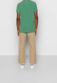 T-shirt en coton vert à manches courtes, associé à un pantalon beige clair. Pieds dans des baskets blanches avec des accents verts. Coupe simple et décontractée.