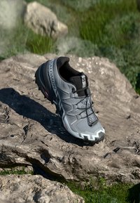 Chaussure de trail running grise avec un dessus texturé, des lacets noirs et un motif de semelle audacieux. Marquage prominent sur le côté. Placée sur une surface rocheuse.