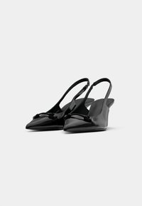 Um par de sapatos de salto alto de couro envernizado preto com pontas finas e detalhe de laço decorativo em fundo branco.