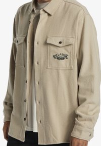 Beige fleece button-up shirt with two front pockets, embroidered "Billabong" logo, and brown buttons; soft texture and relaxed fit.