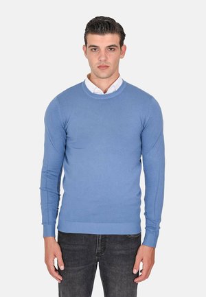 Maglione a maniche lunghe di colore azzurro chiaro, con scollatura rotonda, design aderente e polsini e orlo a coste. Indossato sopra una camicia bianca con colletto.