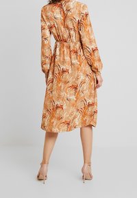 Robe florale beige avec motifs orange et bordeaux, manches longues, taille élastique, et longueur jusqu'aux chevilles. Portée avec des talons rose clair.