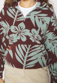 Polarowy sweter z ćwiertkowym zamkiem, w kolorze bordowym z kontrastowym, turkusowym wzorem roślinnym. Zawiera kieszeń z przodu oraz fakturowany materiał.