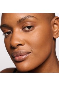 Primer plano de un rostro de piel marrón suave, con labios llenos, un brillo sutil y un delicado aro de plata en la nariz. Una iluminación natural suave realza los rasgos.